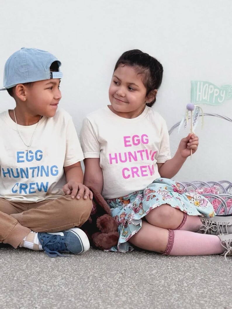 May include: Two children wearing matching cream-colored t-shirts with the text "EGG HUNTING CREW" in blue and pink. One child wears a blue baseball cap and brown pants. The other child wears a floral skirt and pink socks.