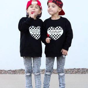 May include: Two children wearing black sweatshirts with a heart-shaped checkered design and the text "Quit Playing Games With My Heart." They wear acid wash jeans, white sneakers, and baseball caps. One cap is red, the other has a lip print.