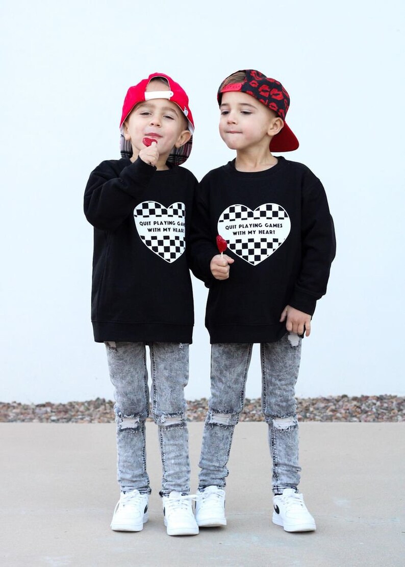 May include: Two children wearing black sweatshirts with a heart-shaped checkered design and the text "Quit Playing Games With My Heart." They wear acid wash jeans, white sneakers, and baseball caps. One cap is red, the other has a lip print.