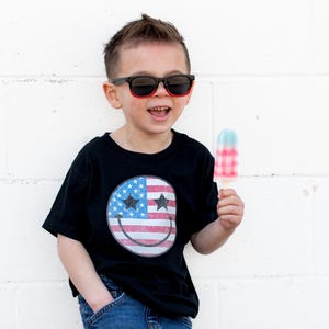 Op de afbeelding: Een kind met een zwarte zonnebril en een zwart t-shirt met een smiley-ontwerp van de Amerikaanse vlag, met een rood, wit en blauw ijsje. De persoon lacht voor een witte bakstenen muur.