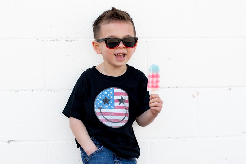 Op de afbeelding: Een kind met een zwarte zonnebril en een zwart t-shirt met een smiley-ontwerp van de Amerikaanse vlag, met een rood, wit en blauw ijsje. De persoon lacht voor een witte bakstenen muur.