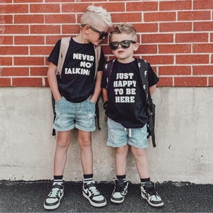 May include: Two black t-shirts with white text. The left shirt reads NEVER NO TALKING and the right shirt reads JUST HAPPY TO BE HERE. Both are paired with light blue distressed denim shorts, black and white sneakers, and black sunglasses.