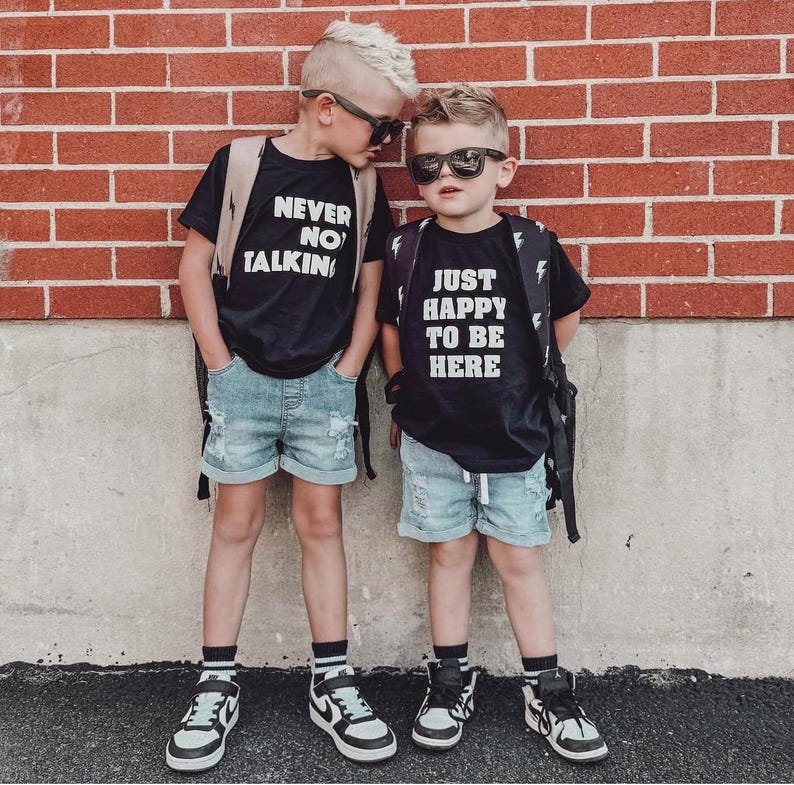 May include: Two black t-shirts with white text. The left shirt reads NEVER NO TALKING and the right shirt reads JUST HAPPY TO BE HERE. Both are paired with light blue distressed denim shorts, black and white sneakers, and black sunglasses.