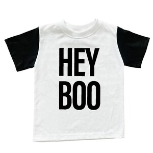 May include: White t-shirt with black sleeves. The words "HEY BOO" are printed in bold black letters on the front of the shirt. The shirt is likely for children and is suitable for Halloween.