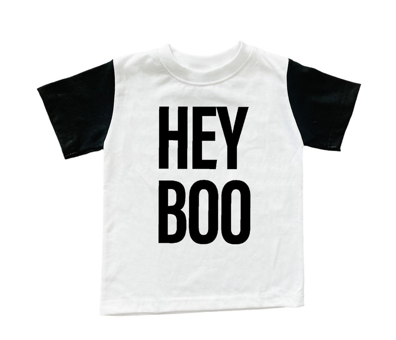 May include: White t-shirt with black sleeves. The words "HEY BOO" are printed in bold black letters on the front of the shirt. The shirt is likely for children and is suitable for Halloween.
