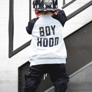 Könnte beinhalten: Ein Kind trägt eine schwarze und weiße Baseballkappe mit Blitzgrafiken und dem Text "REBEL BRO". Das Kind trägt außerdem einen grau-schwarzen Sweatshirt mit dem Aufdruck "BOY HOOD" auf dem Rücken, eine schwarze Hose und graue High-Top-Sneaker.