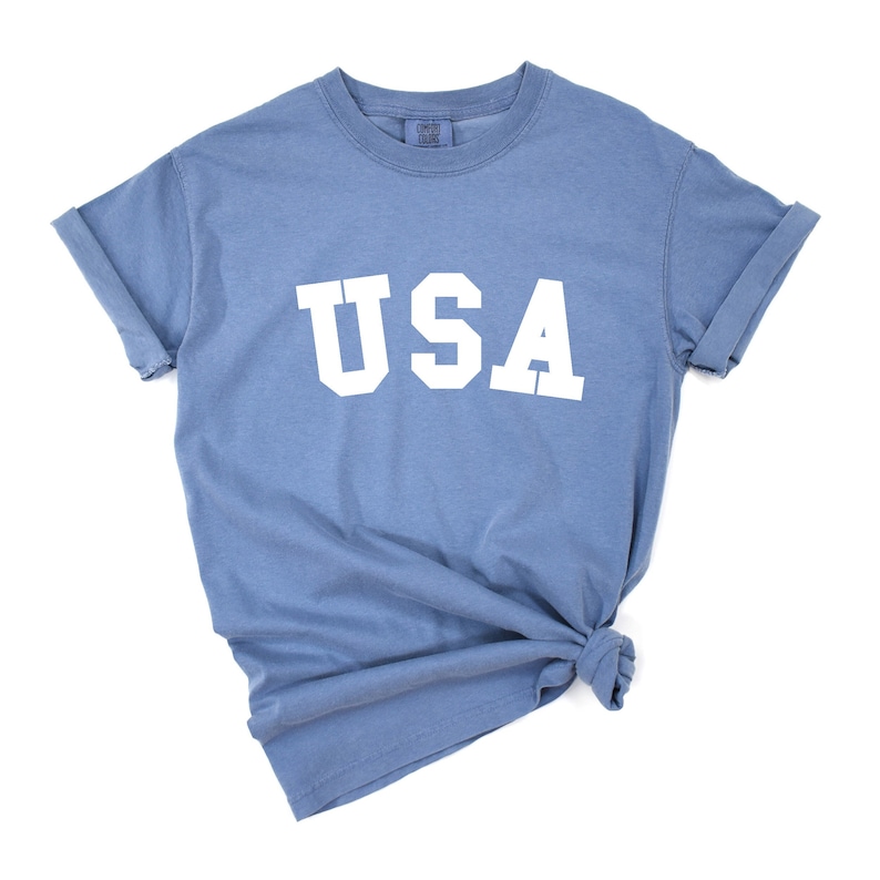 以下が含まれることがあります： 前面に白い文字で「USA」がプリントされた青いTシャツです。