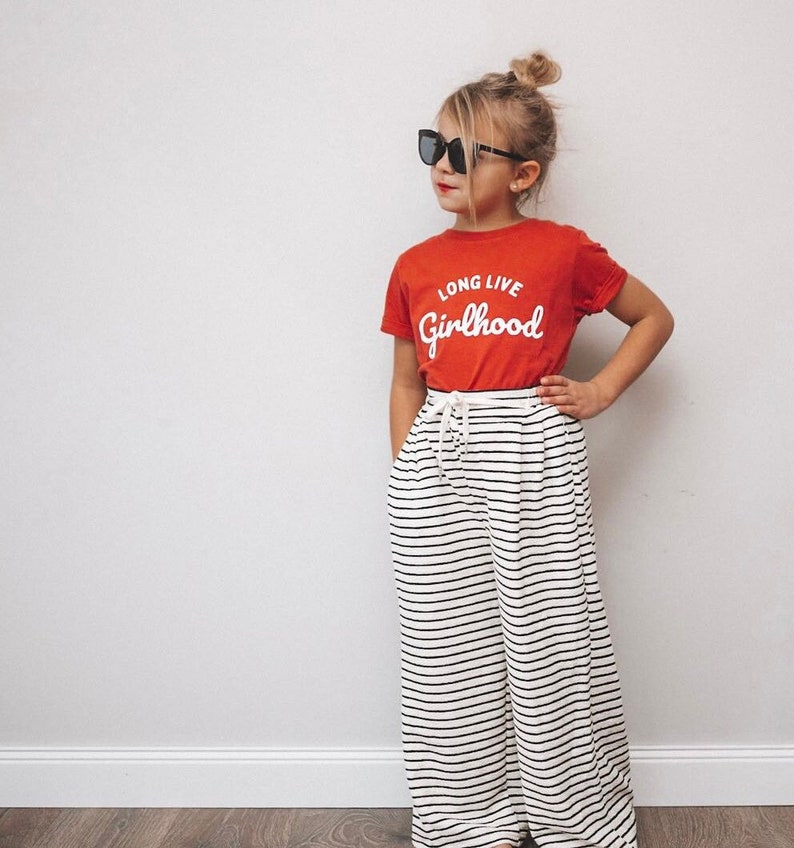 May include: A young person wearing a red t-shirt with the words "LONG LIVE Girlhood" in white script, paired with black and white striped wide-leg pants. The person is also wearing black sunglasses.