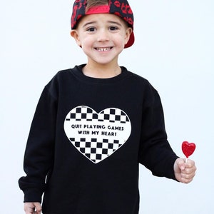 May include: A black sweatshirt with a white heart-shaped design featuring a checkerboard pattern and the text "QUIT PLAYING GAMES WITH MY HEART." The child is wearing a red and black baseball cap and holding a red heart-shaped lollipop.