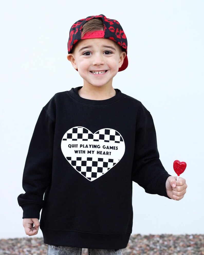 May include: A black sweatshirt with a white heart-shaped design featuring a checkerboard pattern and the text "QUIT PLAYING GAMES WITH MY HEART." The child is wearing a red and black baseball cap and holding a red heart-shaped lollipop.