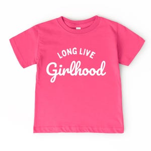 May include: A bright pink t-shirt with the words "LONG LIVE Girlhood" in white script. The shirt has short sleeves and a crew neck. The text is centered on the front of the shirt.