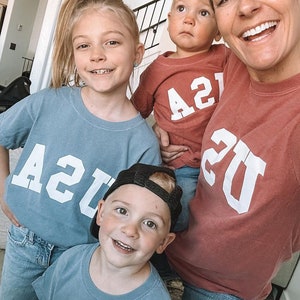 以下が含まれることがあります： 4人家族が、白で「21」とプリントされた、青と赤のマッチングTシャツを着ています。Tシャツは、ヴィンテージな大学のスタイルです。