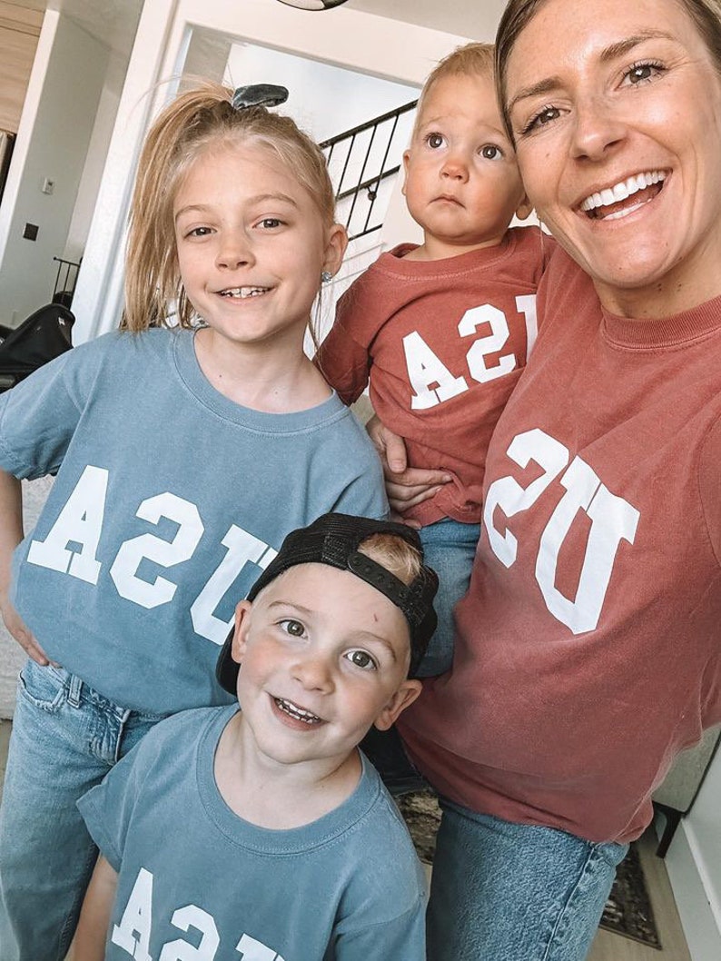 以下が含まれることがあります： 4人家族が、白で「21」とプリントされた、青と赤のマッチングTシャツを着ています。Tシャツは、ヴィンテージな大学のスタイルです。