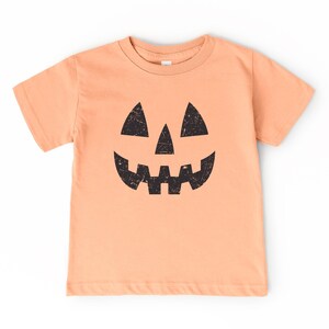 May include: A peach-colored t-shirt featuring a black jack-o'-lantern face. The design includes triangular eyes, a triangular nose, and a wide, toothy grin. The shirt is short-sleeved and suitable for Halloween.