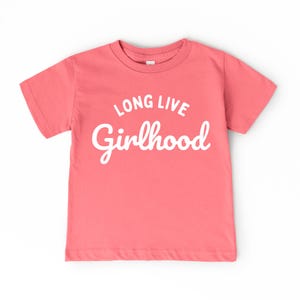 May include: A coral-colored t-shirt with the words "LONG LIVE Girlhood" in white script. The shirt has short sleeves and a crew neck. The text is centered on the front of the shirt.