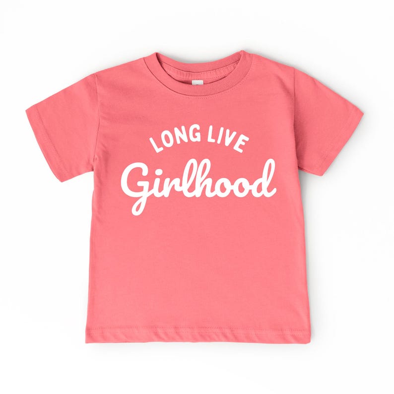 May include: A coral-colored t-shirt with the words "LONG LIVE Girlhood" in white script. The shirt has short sleeves and a crew neck. The text is centered on the front of the shirt.