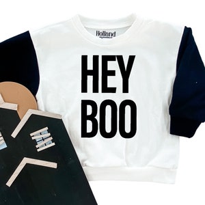 May include: White sweatshirt with black sleeves and the text "HEY BOO" in large black letters. The brand name "Holland" is printed on the upper center. The sweatshirt is a Halloween-themed garment.