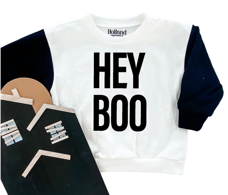 May include: White sweatshirt with black sleeves and the text "HEY BOO" in large black letters. The brand name "Holland" is printed on the upper center. The sweatshirt is a Halloween-themed garment.