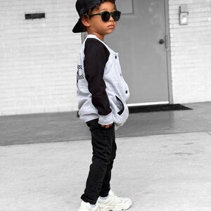 Op de afbeelding: Een jong persoon draagt een zwarte baseballpet, zonnebril, een grijs-zwarte varsity jack, zwarte jeans en witte sneakers. De jas heeft een geruit ontwerp op de rug.