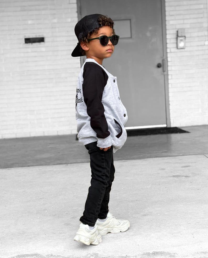 Op de afbeelding: Een jong persoon draagt een zwarte baseballpet, zonnebril, een grijs-zwarte varsity jack, zwarte jeans en witte sneakers. De jas heeft een geruit ontwerp op de rug.