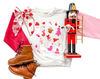 Può includere: Una felpa bianca a maniche lunghe con grafiche a tema natalizio, tra cui Babbo Natale, renne e bastoncini di zucchero. Un fiocco rosso e bianco, uno schiaccianoci di legno e stivali di pelle marrone completano l'arrangiamento festivo.