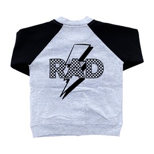Op de afbeelding: Een grijs raglan-stijl sweatshirt met zwarte mouwen. De achterkant van het kledingstuk heeft het woord "RAD" in een geblokt patroon, overliggend met een zwarte bliksem.