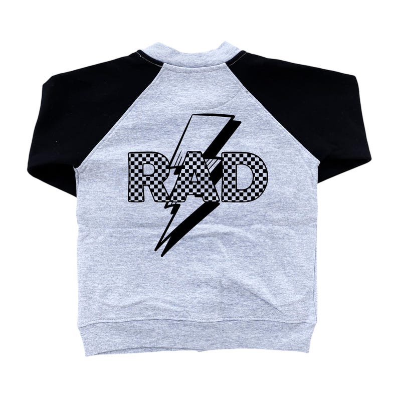 Op de afbeelding: Een grijs raglan-stijl sweatshirt met zwarte mouwen. De achterkant van het kledingstuk heeft het woord "RAD" in een geblokt patroon, overliggend met een zwarte bliksem.