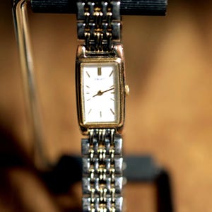 May include: A rectangular Seiko wristwatch featuring a white dial with gold-tone hour markers and hands. The watch case is gold-tone, and the metal link bracelet alternates between silver-tone and gold-tone links, creating a two-tone appearance.