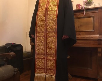 Pode incluir: Um homem vestindo uma túnica preta com uma estola longa bordada em vermelho e dourado. A estola tem franjas na parte inferior e apresenta padrões intrincados.