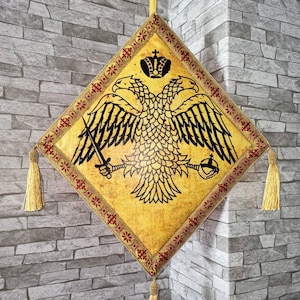 May include: A gold and red embroidered fabric square with a black double-headed eagle design. The eagle has a crown on its head and is holding a sword and a scepter. The fabric has tassels on each corner.