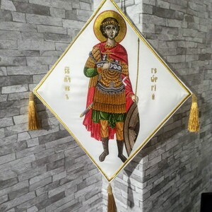 May include: A white diamond-shaped embroidered fabric wall hanging with a gold tassel at each corner. The center of the fabric features a colorful embroidered image of a person in armor holding a sword and shield. The text "Γ'S Γ'P Γ'I" is embroidered in the bottom right corner.