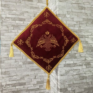 May include: A red velvet square cushion with gold embroidery. The cushion features a two-headed eagle with a crown and a cross. The cushion has gold tassels on each corner and a gold cord hanging from the top.