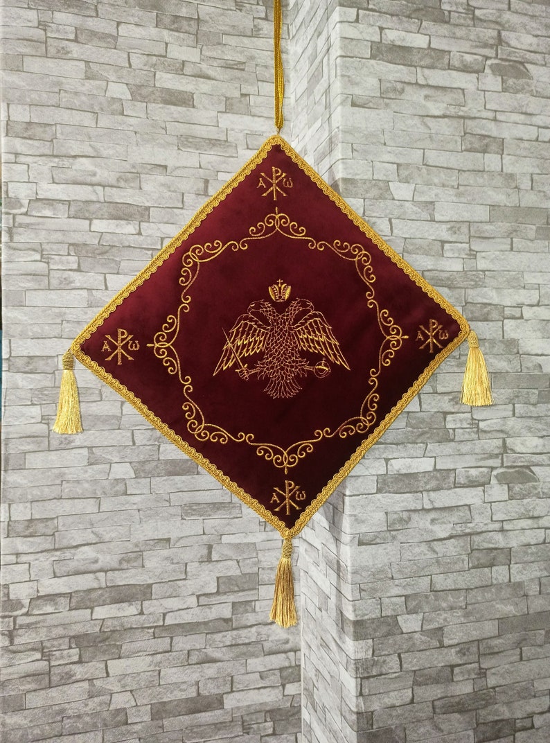 May include: A red velvet square cushion with gold embroidery. The cushion features a two-headed eagle with a crown and a cross. The cushion has gold tassels on each corner and a gold cord hanging from the top.