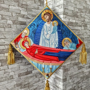 May include: A diamond-shaped embroidered wall hanging with a religious scene depicting Jesus, Mary, and Joseph. The hanging has gold tassels on each corner and is surrounded by a blue border.