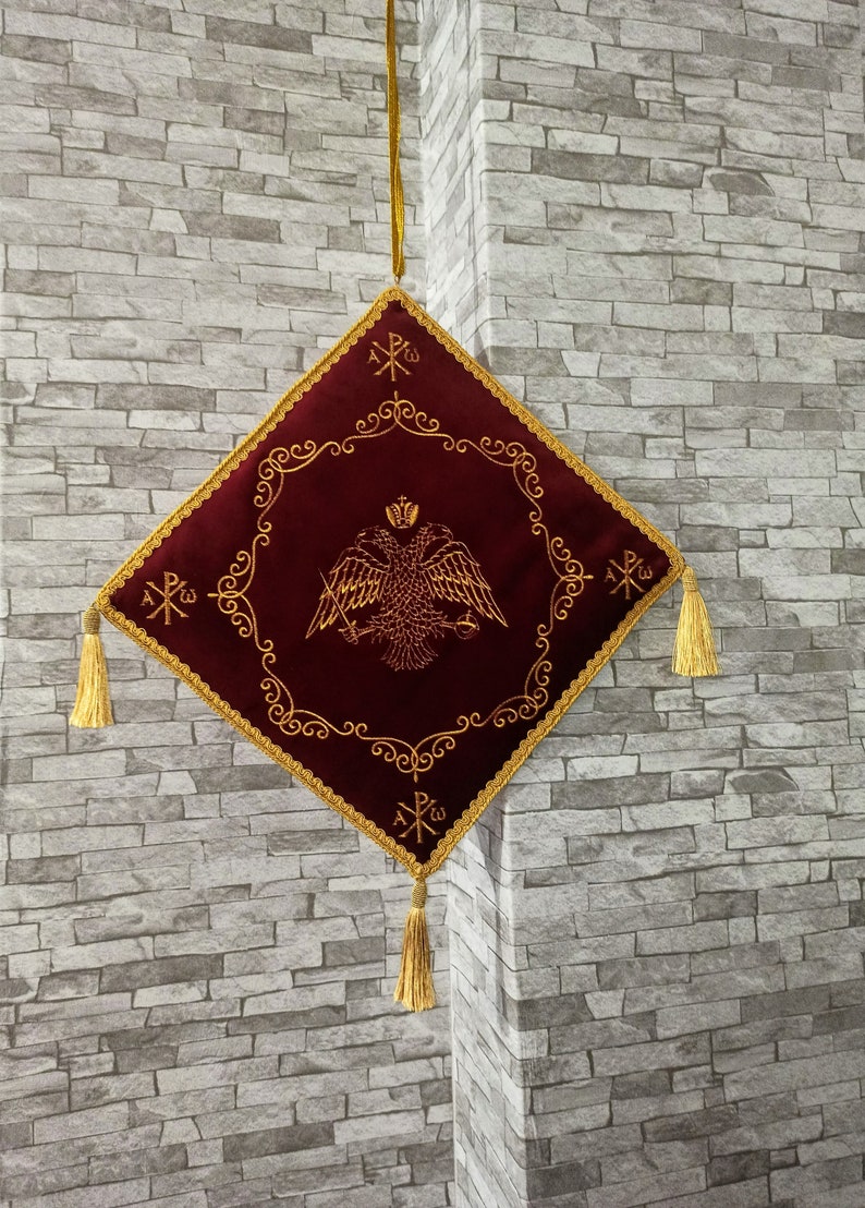 May include: A burgundy velvet square cushion with gold embroidery. The embroidery features a double-headed eagle with a crown, swords, and the Greek letters 'XP'. The cushion has gold tassels on each corner.