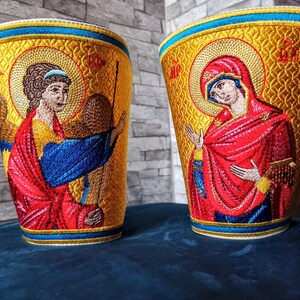 Może przedstawiać: Dwa haftowane pokrowce z religijną ikonografią. Pokrowce są złote z niebieskimi i czerwonymi akcentami. Jeden pokrowiec przedstawia postać kobiety w czerwonych szatach, drugi przedstawia postać mężczyzny w niebieskich i czerwonych szatach.