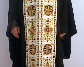 Pode incluir: Uma veste litúrgica branca com bordados dourados e vermelhos. A veste apresenta um padrão repetido de cruzes e círculos. A veste é usada por uma pessoa com uma túnica preta.