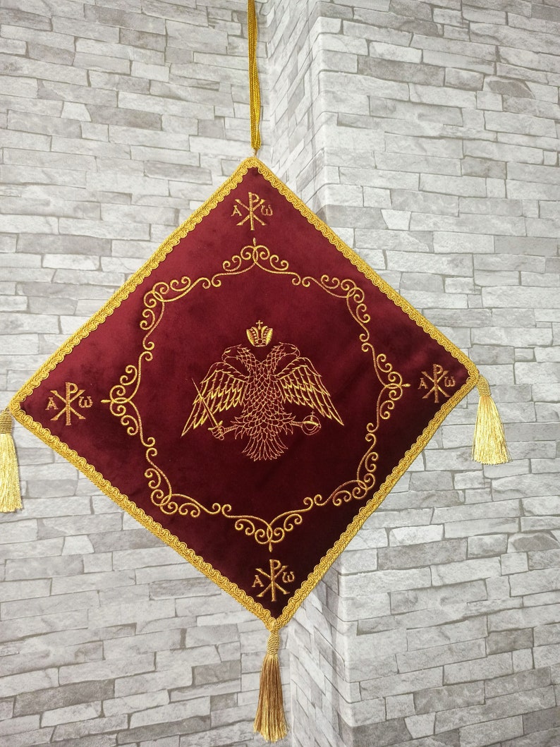 May include: A red velvet square cushion with gold embroidered trim and tassels. The center features a gold embroidered double-headed eagle with a crown and a cross. The text "A&Omicron;&Omega;" is embroidered in the corners.