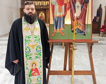 Pode incluir: Um homem vestindo uma batina preta com uma estola bordada em branco e dourado. Ele está em pé na frente de um cavalete de madeira com um ícone religioso retratando duas figuras. O ícone tem um fundo dourado e as figuras são pintadas em vermelho, azul e branco. O texto no ícone diz "ΟΙ ΑΓΙΟΙ ΠΑΤΕΡΕΣ", que se traduz como "Os Santos Padres".