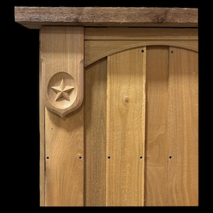 May include: A light brown wooden structure featuring vertical planks and a horizontal top beam. A decorative wooden accent piece is attached to the left side, displaying a carved five-pointed star centered within a shield-shaped frame.