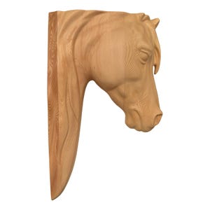 May include: A wooden sculpture of a horse's head and neck, carved from light-colored wood. The sculpture is detailed, showing the horse's mane, eye, and facial features. The piece is designed to be mounted on a wall.