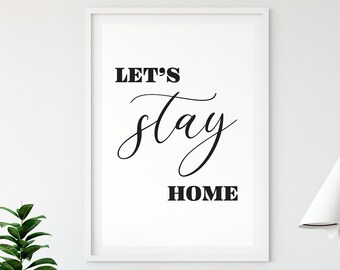 May include: A white framed print with black text that reads "Let's stay home". The print is hanging on a white wall with a wooden chair and a plant in a white pot in the foreground.