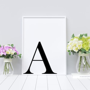 May include: A black and white printable wall art print with the letter A in a bold, sans-serif font. The print is framed in a white frame and is displayed on a white wall with two vases of flowers on either side.