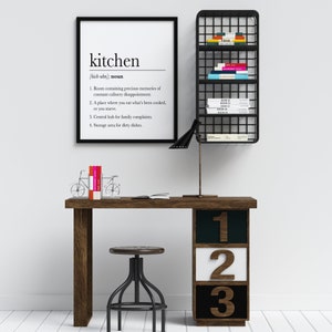 May include: A black and white framed print with the definition of the word "kitchen" hanging on a white wall. A black metal shelf with books on it is mounted on the wall. A wooden desk with a black lamp, a stool, and a bookend shaped like a bicycle is in front of the wall. The desk has three drawers with the numbers 1, 2, and 3 on them.