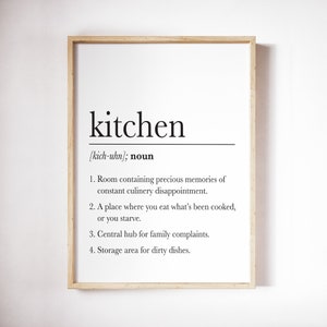 May include: A framed print with a humorous definition of the word "kitchen" in black text on a white background. The definition includes phrases like "precious memories", "culinary disappointment", "family complaints", and "dirty dishes".