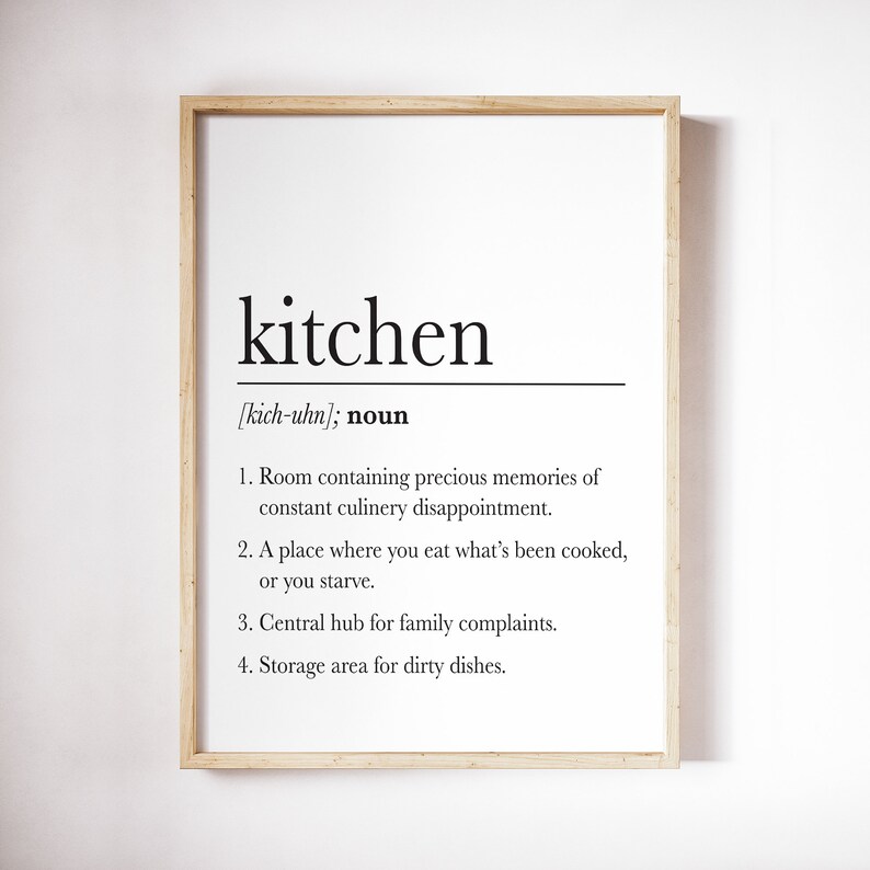 May include: A framed print with a humorous definition of the word "kitchen" in black text on a white background. The definition includes phrases like "precious memories", "culinary disappointment", "family complaints", and "dirty dishes".