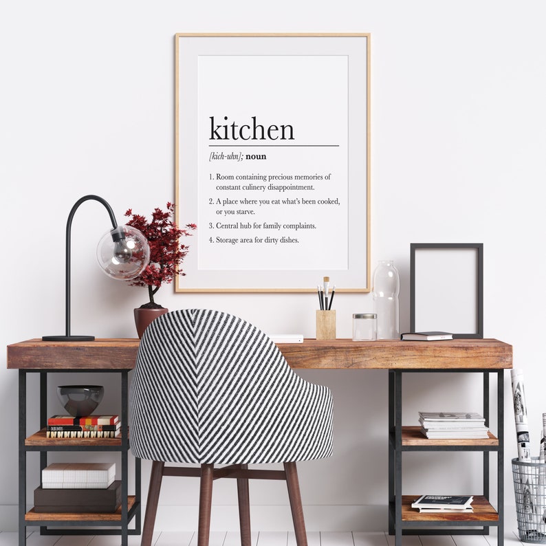 May include: A framed print with a black and white definition of the word "kitchen" with a humorous description of the room. The print is hanging on a white wall above a wooden desk with a black metal frame. A black lamp with a clear glass globe is on the desk next to a small red plant. A black and white striped chair is in front of the desk.
