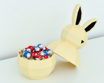 May include: A yellow papercraft bunny with black ears holding a yellow papercraft egg filled with red and blue foil-wrapped chocolate eggs.