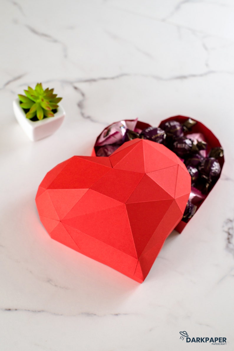 May include: A red paper heart-shaped box with a geometric design. The box is open and filled with small, dark chocolate candies.