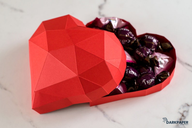 May include: A red paper heart-shaped box with a geometric design, open to reveal a collection of dark chocolate truffles wrapped in silver foil.